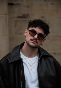 Schwarze Lederjacke, weißes T-Shirt, goldene Gliederkette, rot getönte Sonnenbrille, kurze lockige Haare, stehend vor einem strukturierten Stein-Hintergrund.