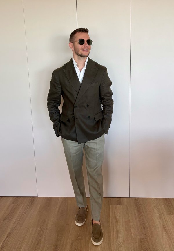 Mann mit Sonnenbrille, dunklem Doppelreiher-Blazer, weißem Hemd, hellen Hosen und braunen Schuhen steht mit den Händen in den Taschen drinnen.