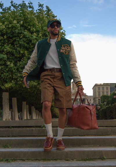 Chaqueta varsity verde y beige con logo, camiseta blanca, pantalones cortos marrones, bolsa de cuero marrón, calcetines blancos, zapatos de plataforma marrones. Gorra verde y gafas de sol.