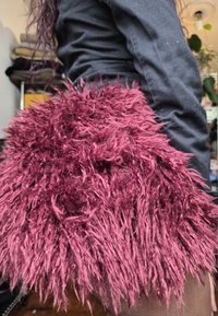 Manga de pelagem sintética em bordeaux profundo, com fibras longas e desgrenhadas. Acompanhada por uma camisa justa e escura. Itens ao fundo desfocados.