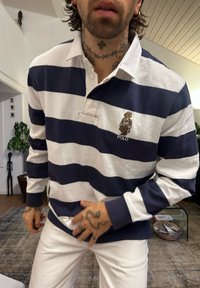 Polo a righe blu navy e bianche con colletto, con un logo a forma di orso cucito sul petto a sinistra. Indossato con pantaloni bianchi, mostrando braccia tatuate.