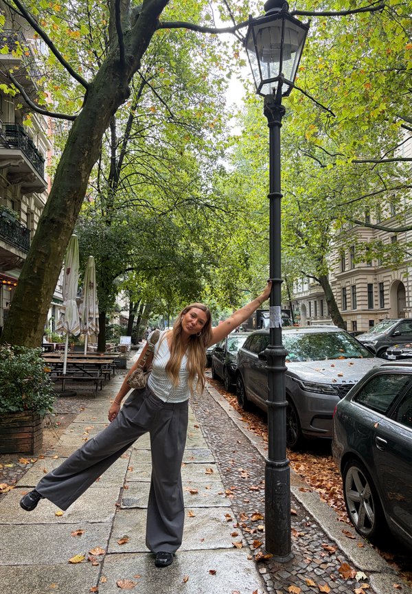 Jeune femme debout sur une jambe tenant un lampadaire sur un trottoir urbain bordé d’arbres avec des voitures garées et des feuilles d’automne.