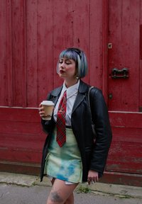 Modèle portant une veste en cuir noire, une chemise blanche, une cravate à carreaux rouges et une jupe tie-dye pastel, tenant une tasse de café. L'arrière-plan présente un mur rouge.