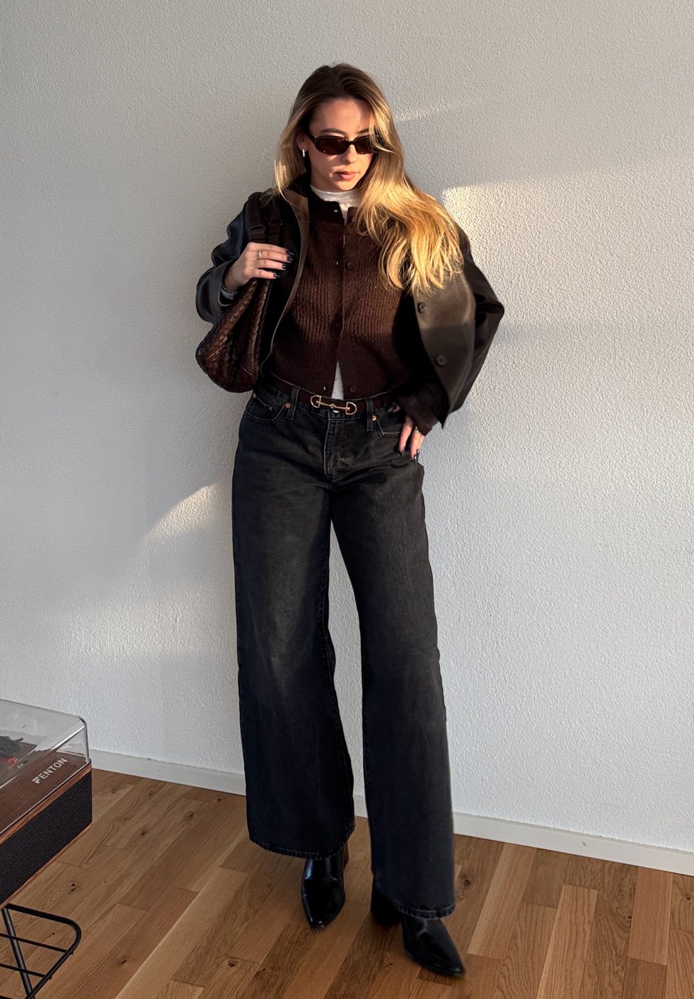 Femme aux longs cheveux blonds portant des lunettes de soleil, un pull marron, une veste en cuir noire, un jean large noir, des bottes noires, et tenant un sac marron à l'intérieur.