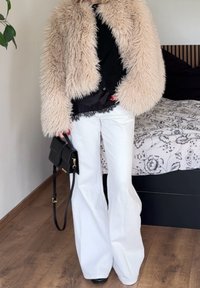 Veste en fausse fourrure beige, haut noir ajusté avec bordure en dentelle, pantalon blanc évasé et petit sac à main noir avec bandoulière.