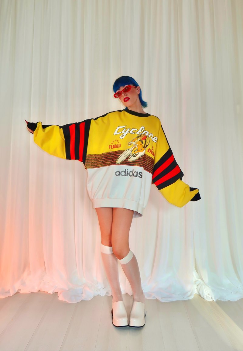 Sweatshirt amarelo oversized com riscas pretas e vermelhas, estampa gráfica, parte inferior branca, combinado com meias brancas até ao joelho e sapatos de salto.