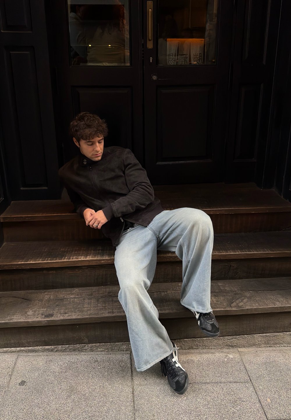 Fekete cipzáras dzseki, laza, világoskék, enyhén bővülő szárú farmer, és fekete sportcipő. Egy sötét ajtó előtt, fából készült lépcsőkön ül.