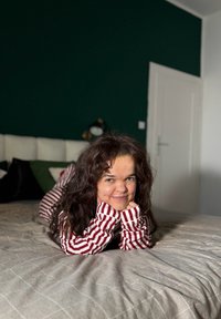 Femme aux cheveux bruns bouclés portant un pyjama rouge à rayures blanches, allongée sur le ventre sur un lit dans une chambre aux murs verts foncés.