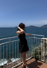 Vestido preto sem mangas com um corpete justo, usado por uma pessoa em pé num balcão a olhar para as ondas azuis do oceano e colinas distantes.