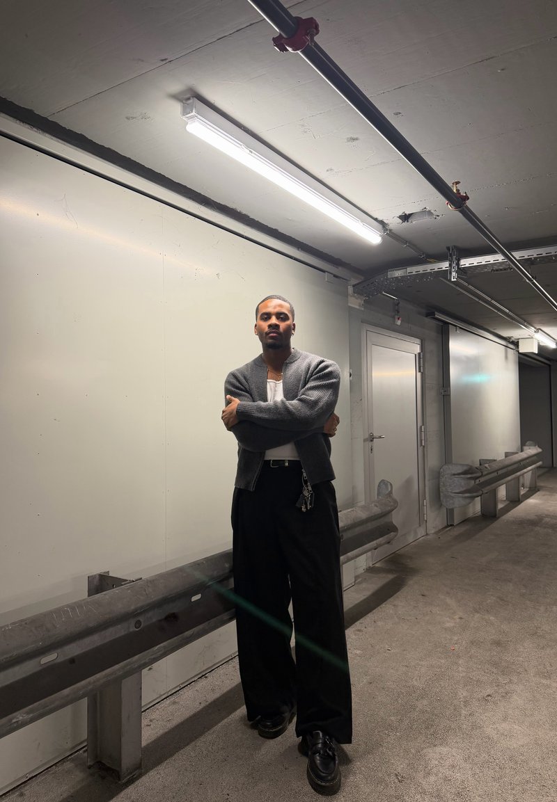 Soft, grey cardigan over a white shirt, paired with loose black trousers and black shoes, in a minimal concrete corridor with overhead lights.