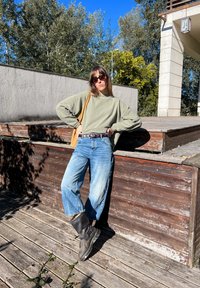 Sweatshirt oversized verde-oliva, jeans largos azuis claros e botas de tornozelo pretas. O fundo apresenta um deck de madeira e árvores.