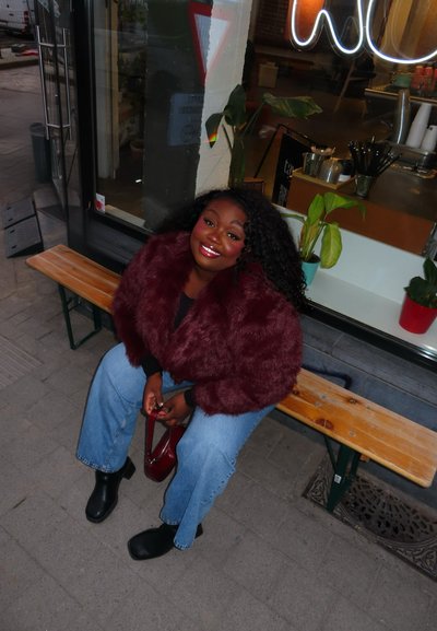 Mujer con cabello rizado que lleva un abrigo de piel burdeos y vaqueros azules, sentada en un banco de madera frente a una cafetería con plantas en macetas en el interior.