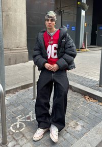 Jeune personne portant un bonnet camouflage, un maillot de sport rouge, une doudoune noire et un pantalon large noir assise sur un porte-vélos en métal sur un trottoir urbain.