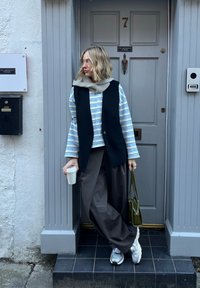 Černá vesta přes modro-bíle pruhované tričko, šedé kalhoty s širokými nohavicemi, drží papírový kelímek. Stříbrné tenisky a zelená taška. Pozadí s texturovanými dveřmi.