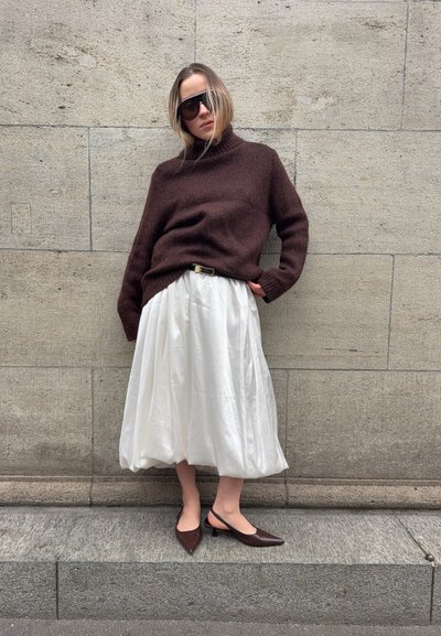 Persona con suéter de cuello alto marrón oscuro, falda midi blanca voluminosa, tacones slingback marrón oscuro y gafas de sol grandes, de pie junto a una pared de piedra.