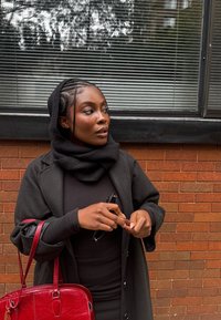 Cappotto di lana nero indossato sopra un vestito nero, sciarpa nera con trama, occhiali da sole in mano, con una borsa rossa in pelle su uno sfondo di muro di mattoni.