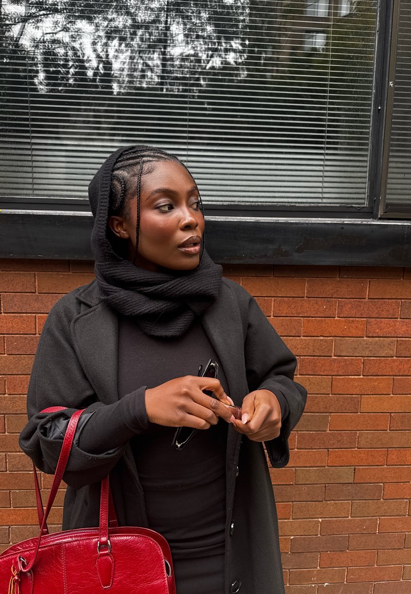 Manteau en laine noire porté sur une robe noire, écharpe noire texturée, tenant des lunettes de soleil, avec un sac à main en cuir rouge sur fond de mur en briques.