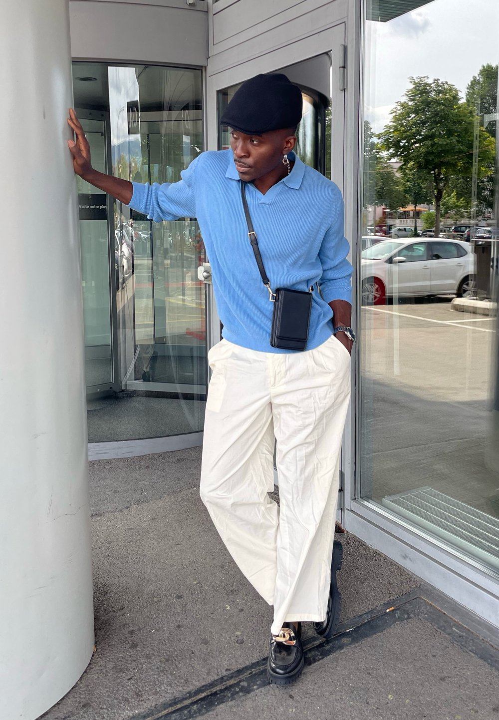 Lyseblå ribbestrikket genser, hvite vide bukser og svarte sko. En svart crossbody veske og caps. Står nær en glassinngang.