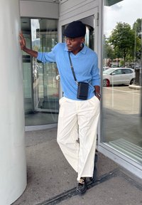 Lyseblå ribbestrikket genser, hvite vide bukser og svarte sko. En svart crossbody veske og caps. Står nær en glassinngang.