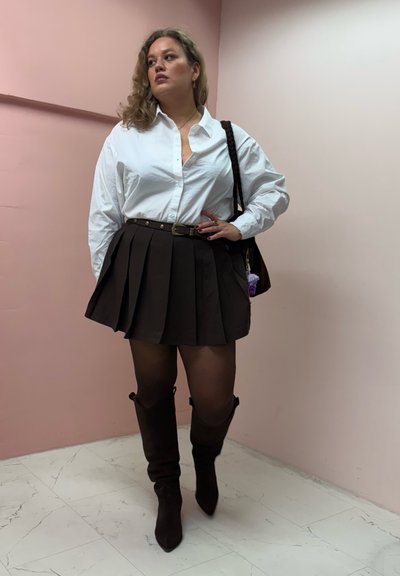 Camisa blanca de botones, falda corta marrón con pliegues y cinturón, medias negras y botas marrones hasta la rodilla. El fondo es una pared rosa claro.