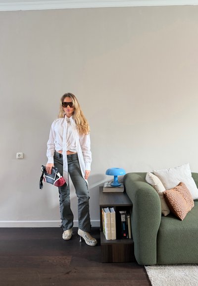 Mujer con camisa blanca, pantalones grises y zapatos de tacón alto, de pie cerca de un sofá verde y una mesa auxiliar con una lámpara azul y libros.