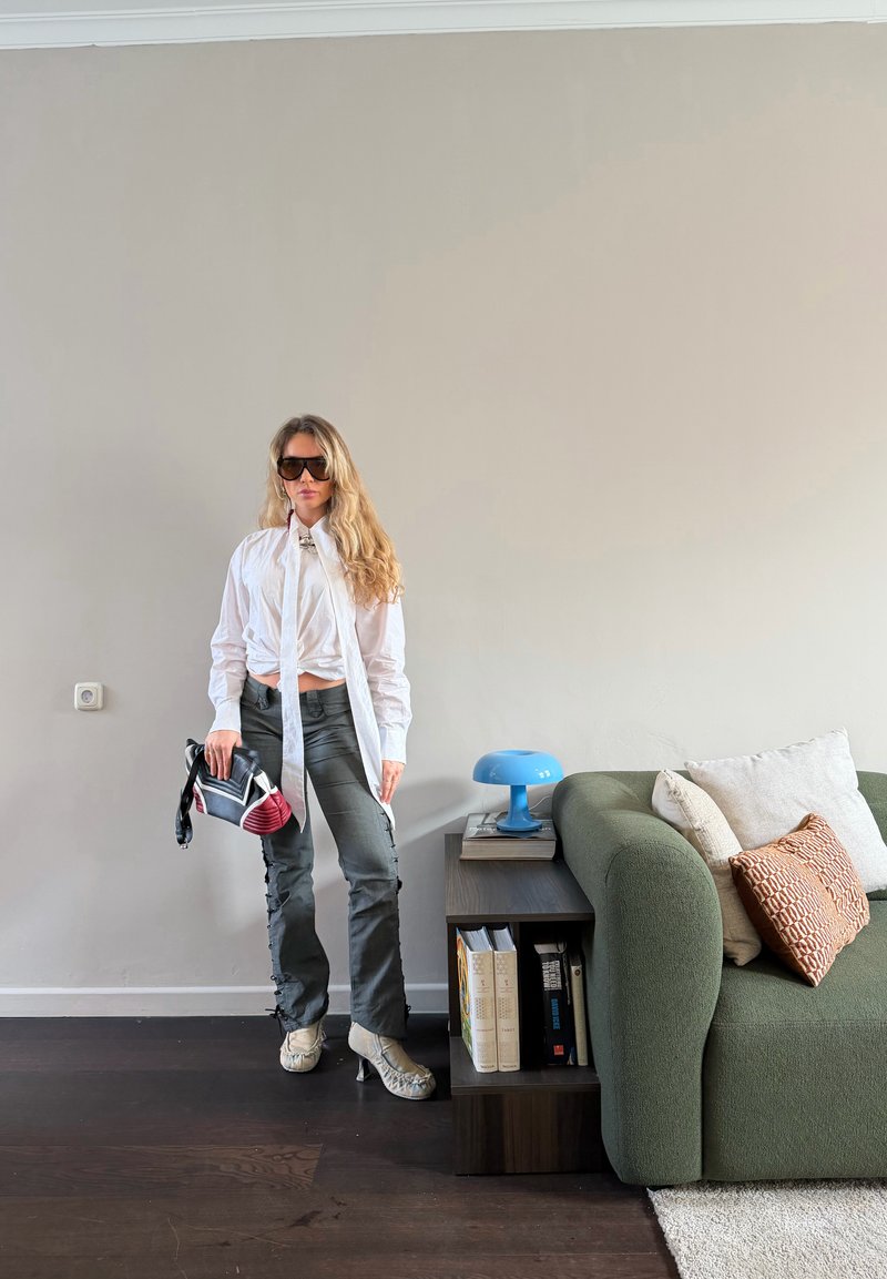 Femme portant une chemise blanche, un pantalon gris et des chaussures à talons hauts se tient près d'un canapé vert et d'une table d'appoint avec une lampe bleue et des livres.