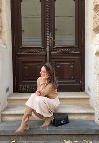 Bež pleteni džemper, krem midi suknja, taupe ante čizme s potpeticama. Crna torba s zlatnim detaljima. Smeđa drvena vrata u pozadini.