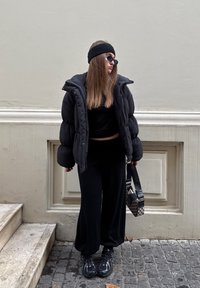 Giacca imbottita nera, top nero corto, pantaloni neri larghi, fascia nera per capelli, occhiali da sole e borsa decorata con tracolle.