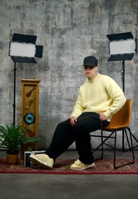 Pull tricoté jaune, pantalon noir ample, baskets beige. Casquette noire, assis sur une chaise en bois devant un mur gris texturé, avec des objets décoratifs.