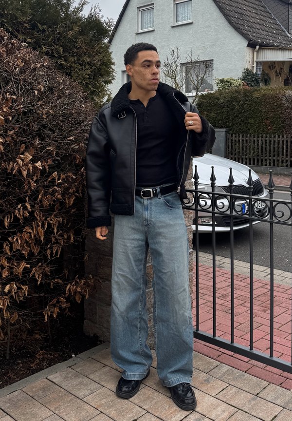 Giovane uomo con giacca di pelle nera, camicia nera, jeans larghi blu e scarpe nere che si trova su un sentiero lastricato vicino a un cancello e un'auto.