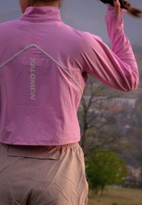 Žmonės nugaros vaizdas dėvint šviesiai violetinę Salomon striukę, lauke gamtos aplinkoje reguliuojant uodegą, aplink medžiai ir tolimoje vietovėje matomi pastatai.