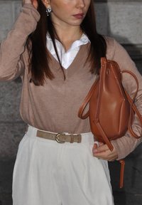 Hellbrauner V-Ausschnitt-Pullover, darunter ein weißes Kragenhemd, beiger Gürtel, hochgeschnittene weiße Hose und eine kleine braune Ledertasche.