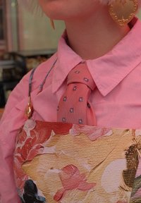 Rosa Hemd mit Rüschensleeves, rot-blauer Blumenbind mit floralem Schürze aus strukturiertem Stoff in lebhaften Farben. Goldene herzförmige Ohrringe.