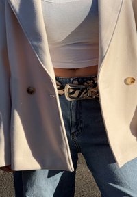 Casaco creme de duas fileiras de botões, top cropped branco canelado, jeans de cintura alta e um cinto estampado com fivela prateada.