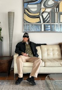 Veste en cuir noire portée sur une chemise vert olive, pantalon beige et baskets noires. Assis sur un canapé en cuir blanc capitonné dans un espace moderne.