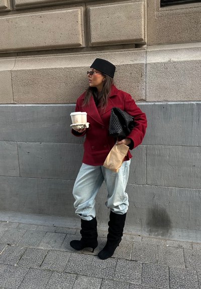 Abrigo rojo de doble botonadura; vaqueros azules claros y holgados; botas negras altas y caídas; sosteniendo una taza de café y un bolso negro tejido, apoyada contra una pared.