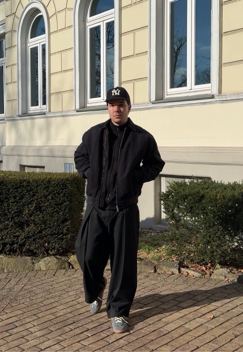 Homme portant une veste noire, un pantalon large noir, des baskets grises et une casquette de baseball noire, marchant sur un chemin en briques à côté d'un bâtiment beige avec de grandes fenêtres.