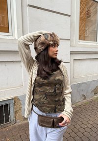 Cardigan a coste crema, top militare verde a camuflaggio, pantaloni della tuta grigi e berretto foderato in pelliccia. Borsa in vita marrone con due tasche.