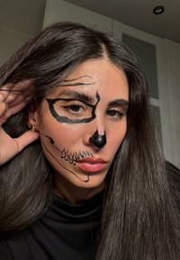 Zwarte gezichtsschilderingen die op een schedel lijken aan de ene kant van het gezicht, met gedurfde lijnen en een gedefinieerde neus, gecombineerd met lang, donker haar.