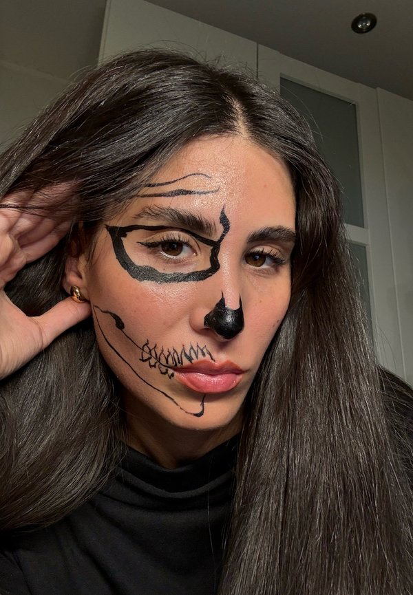 Des motifs de peinture noire sur le visage ressemblant à un crâne sur un côté du visage, avec des lignes audacieuses et un nez défini, associés à de longs cheveux noirs.