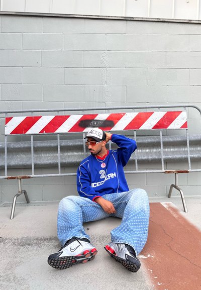 Jersey deportivo azul con acentos rojos, jeans azul claro con un patrón y zapatillas plateadas y negras. Sentado contra una pared gris, con una barrera detrás.