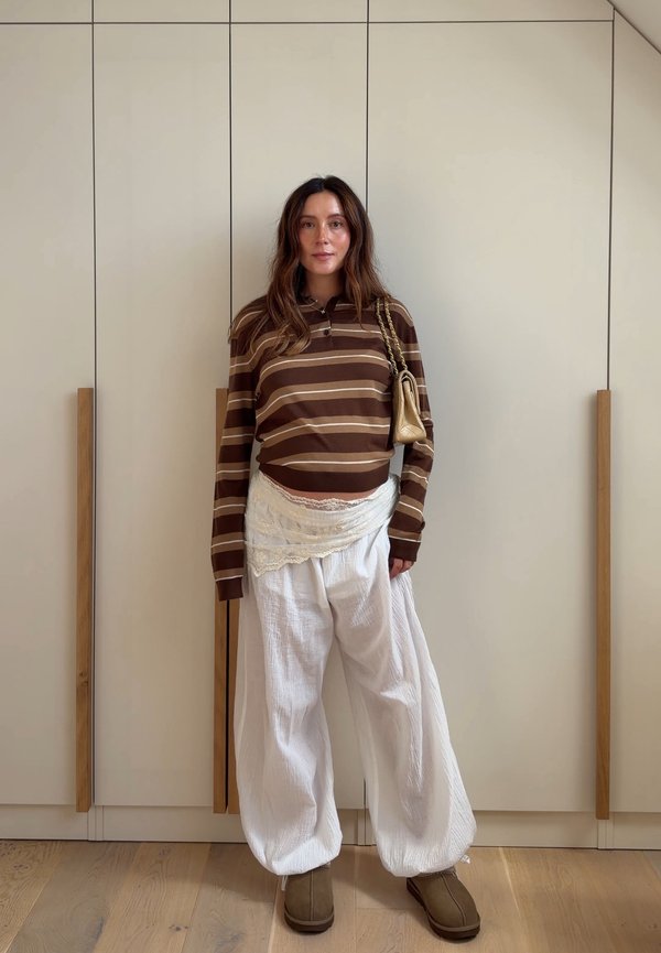 Femme debout à l'intérieur portant une chemise à manches longues rayée marron, un pantalon large blanc, des bottes beige clair et portant un petit sac beige à l'épaule.