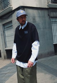 Fekete, túlméretes póló kék "Calvin Klein" logóval, fehér csíkozott ingre rétegezve. Kék csíkozott sapkát és olivazöld nadrágot visel.
