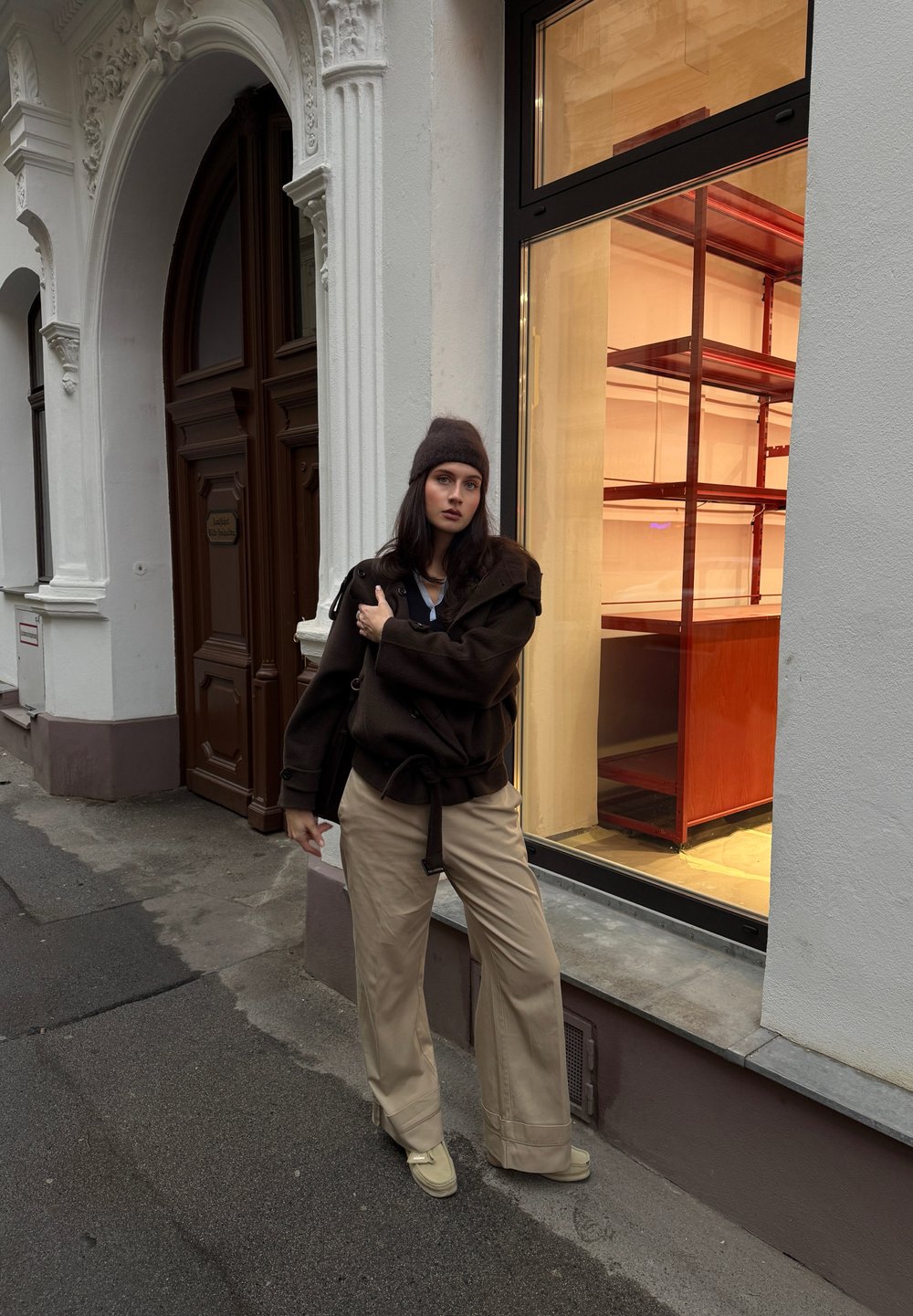 Pull en maille marron oversize avec ceinture, pantalon beige à jambes larges, et chaussures beiges. En arrière-plan, une vitrine de magasin en verre avec des étagères en bois.