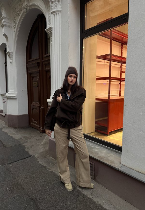 Maglione marrone oversize in maglia con cintura, pantaloni beige a gamba larga e scarpe beige. Lo sfondo mostra una vetrina di un negozio in vetro con scaffali in legno.