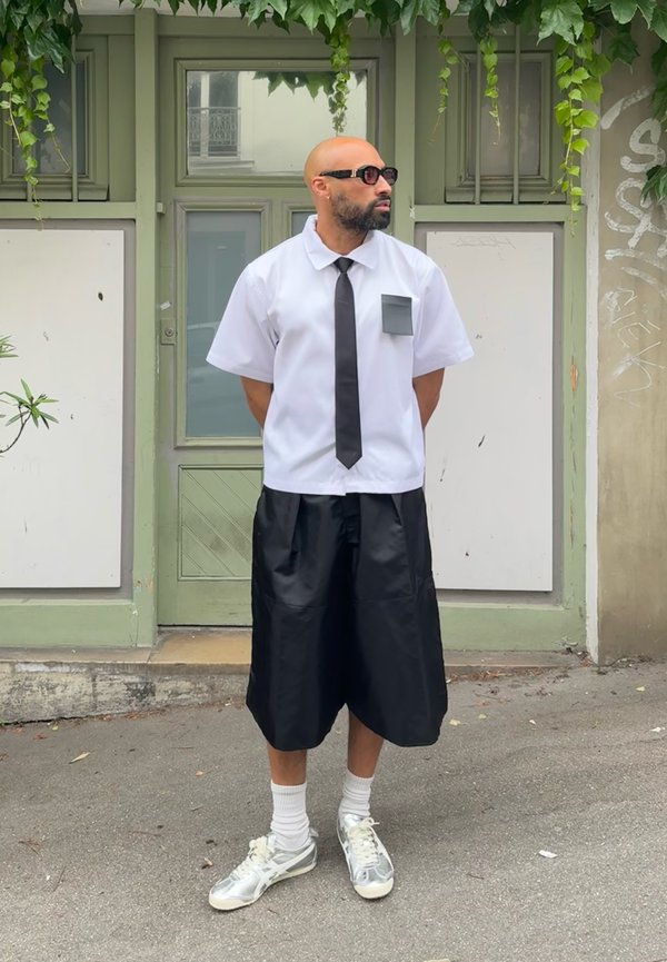 Camicetta bianca a maniche corte con una cravatta nera e tasca, abbinata a pantaloni corti neri fino al ginocchio e sneakers bianche con accenti grigi.