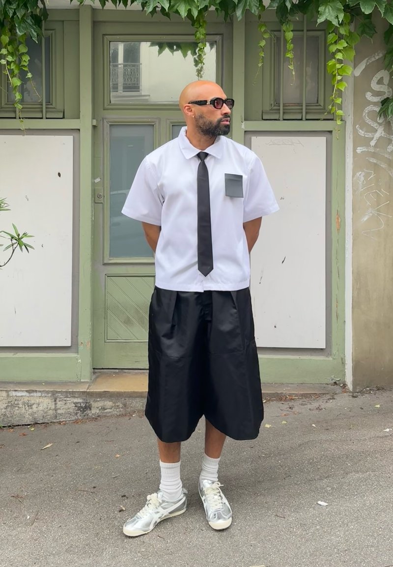 Λευκό μπλουζάκι κοντομάνικο με μαύρη γραβάτα και τσέπη, συνδυασμένο με μαύρες βερμούδες μέχρι το γόνατο και λευκά αθλητικά παπούτσια με γκρι λεπτομέρειες.