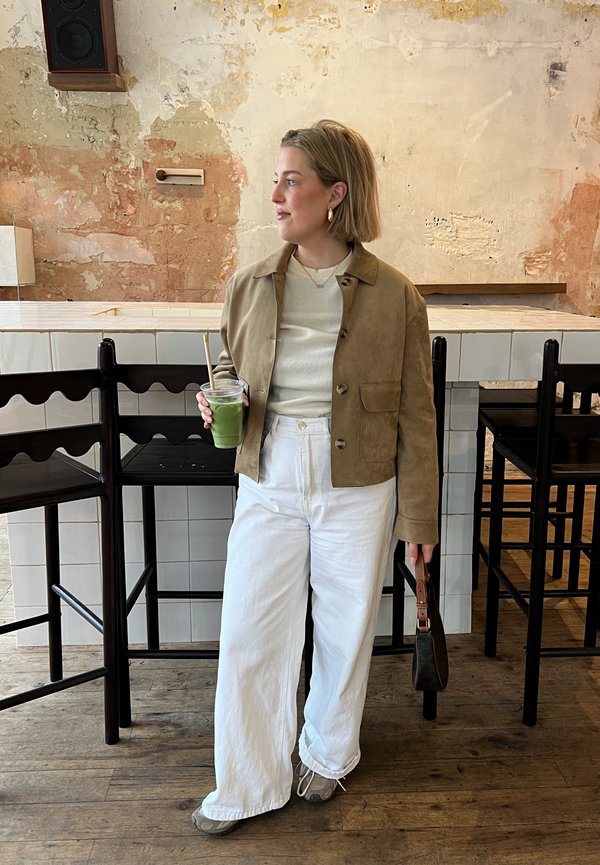 Ženska v bež jakni in belih hlačah drži zelen napitek in majhno torbico, stoji ob barskih stolih v rustikalnem kavarniškem ambientu.