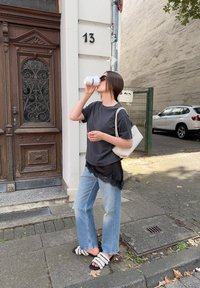 Tricou gri suprapus peste un top negru cu dantelă, blugi de un albastru deschis, poșetă albă și sandale negre. Bea dintr-o cană albă.