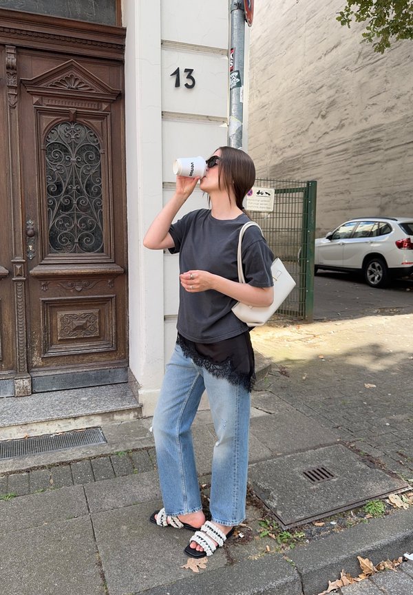 Szary t-shirt nałożony na czarną bluzkę z koronkowym wykończeniem, jasnoniebieskie dżinsy, biała torebka i czarne sandały. Pije z białego kubka.